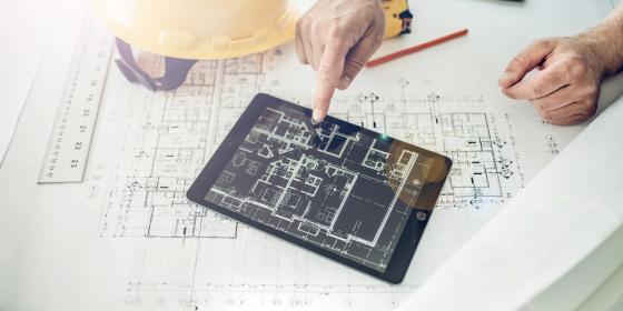 Ein Tablet liegt auf einem ausgerollten Bauplan und wird von einer Hand bedient