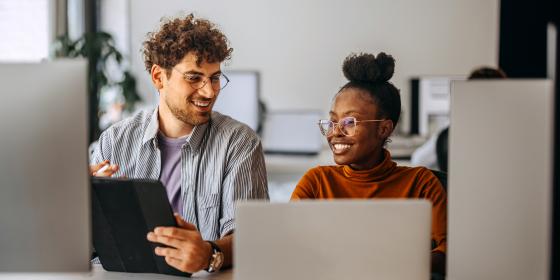 Zwei junge lächelnde Personen sitzen an einem Schreibtisch vor zwei Laptops