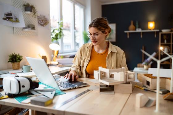 Eine blonde Frau mit Brille sitzt an einem Tisch, auf dem ein Laptop und Baumodell-Teile stehen