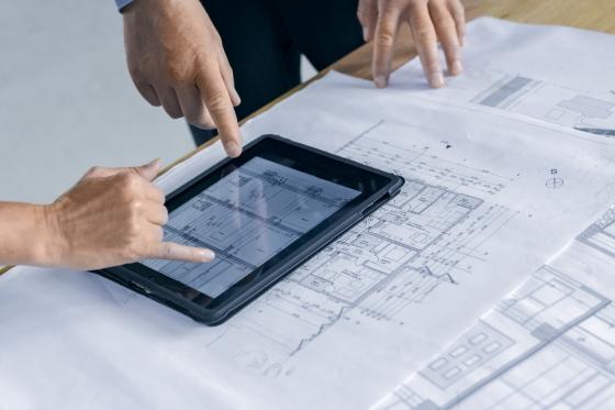 Zwei Finger zeigen auf ein Tablet, das auf einem Tisch mit Bauplänen liegt