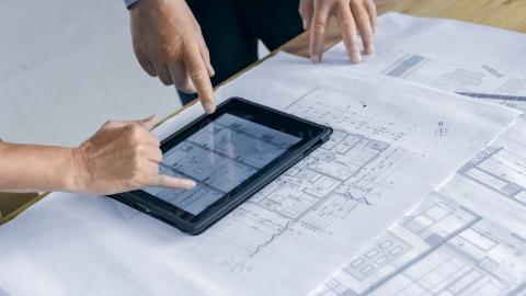 Ein Tablet liegt auf einem ausgerollten Bauplan und wird von einer Hand bedient