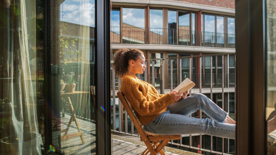 Eine Frau sitzt auf einem Holzstuhl auf einem Balkon und liest ein Buch.