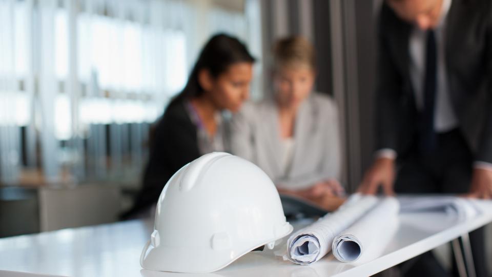Ein Bauhelm liegt mit zusammengerollten Papieren auf einem Tisch, an dem drei Menschen arbeiten